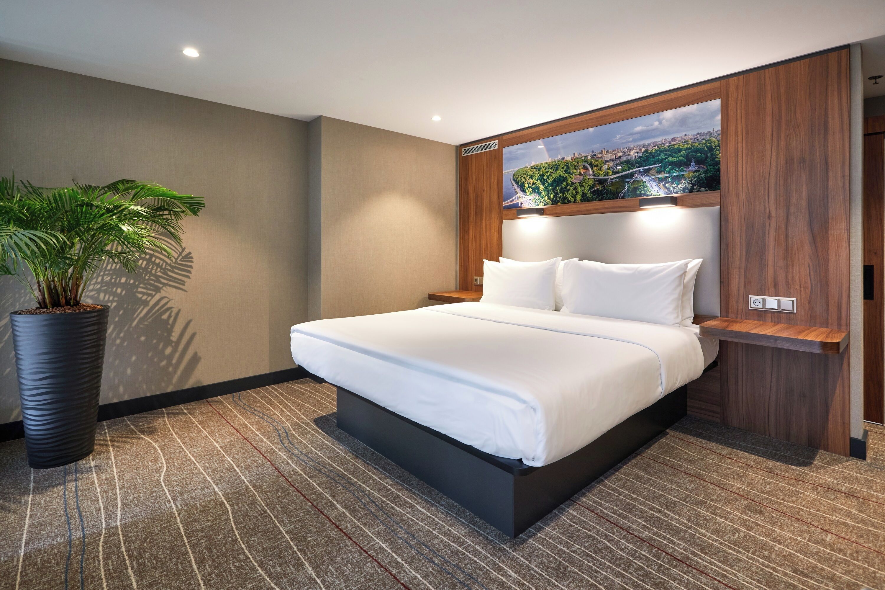 A modern hotel room with a large bed, a potted plant, and a cityscape view through a window.