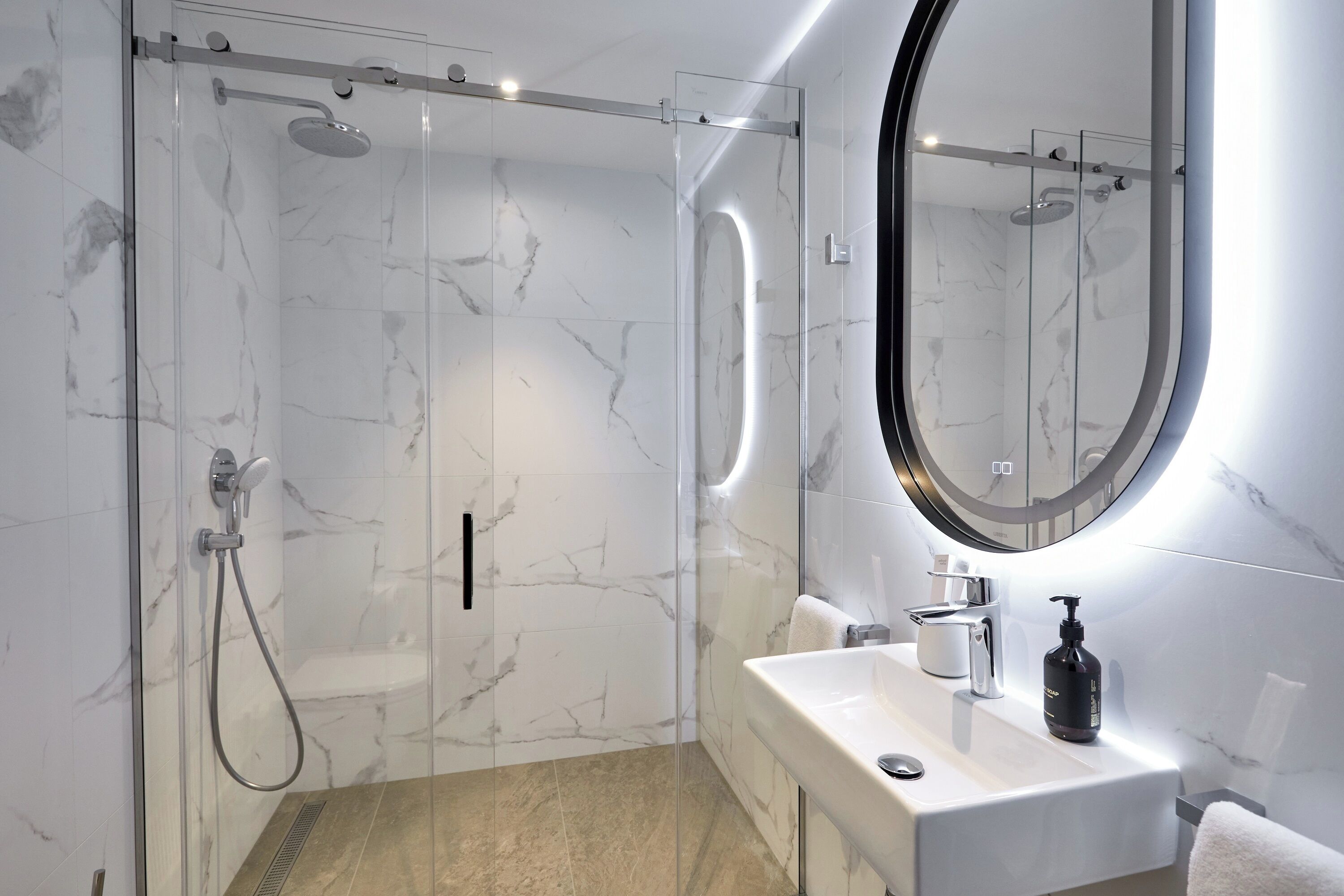 Un baño moderno con ducha acristalada, lavamanos blanco con espejo y paredes de mármol.