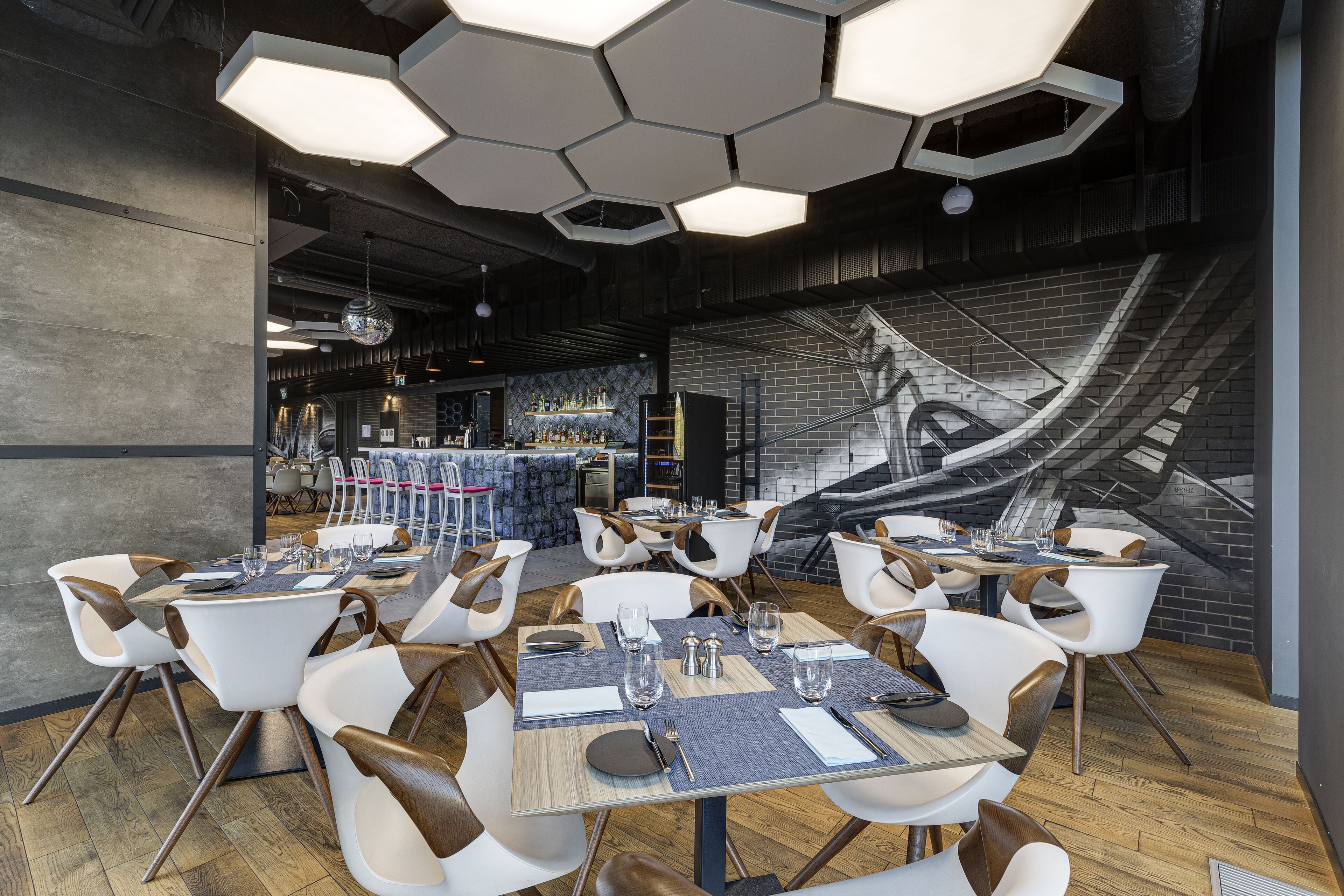 Un restaurant moderne avec des luminaires hexagonaux au plafond, des chaises blanches et marron, et une fresque murale.
