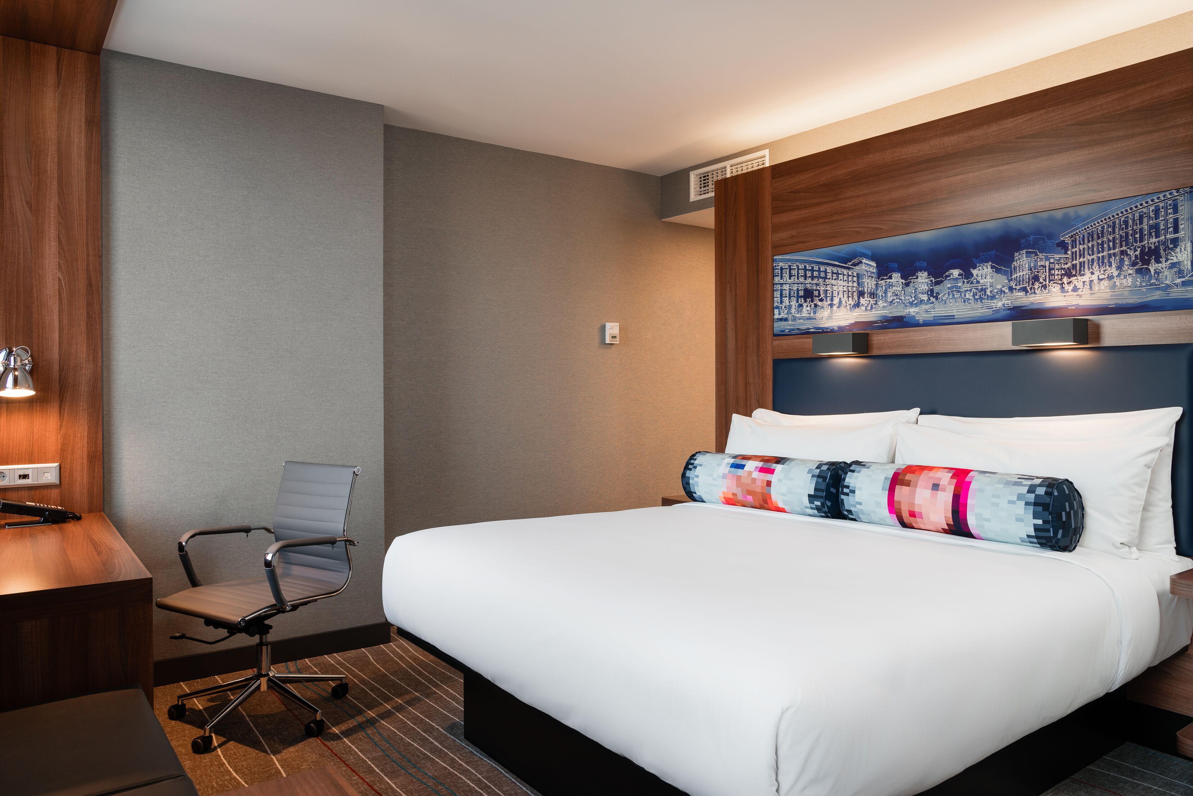 Une chambre d’hôtel avec un grand lit, un bureau, une chaise et une œuvre d’art représentant un paysage urbain au niveau de la tête de lit.