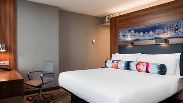 Une chambre d’hôtel avec un grand lit, un bureau, une chaise et une œuvre d’art représentant un paysage urbain au niveau de la tête de lit.