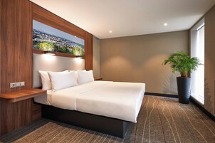 Een moderne hotelkamer met een groot bed, uitzicht op een stadslandschap door een raam, een kamerplant en een houten hoofdeinde.