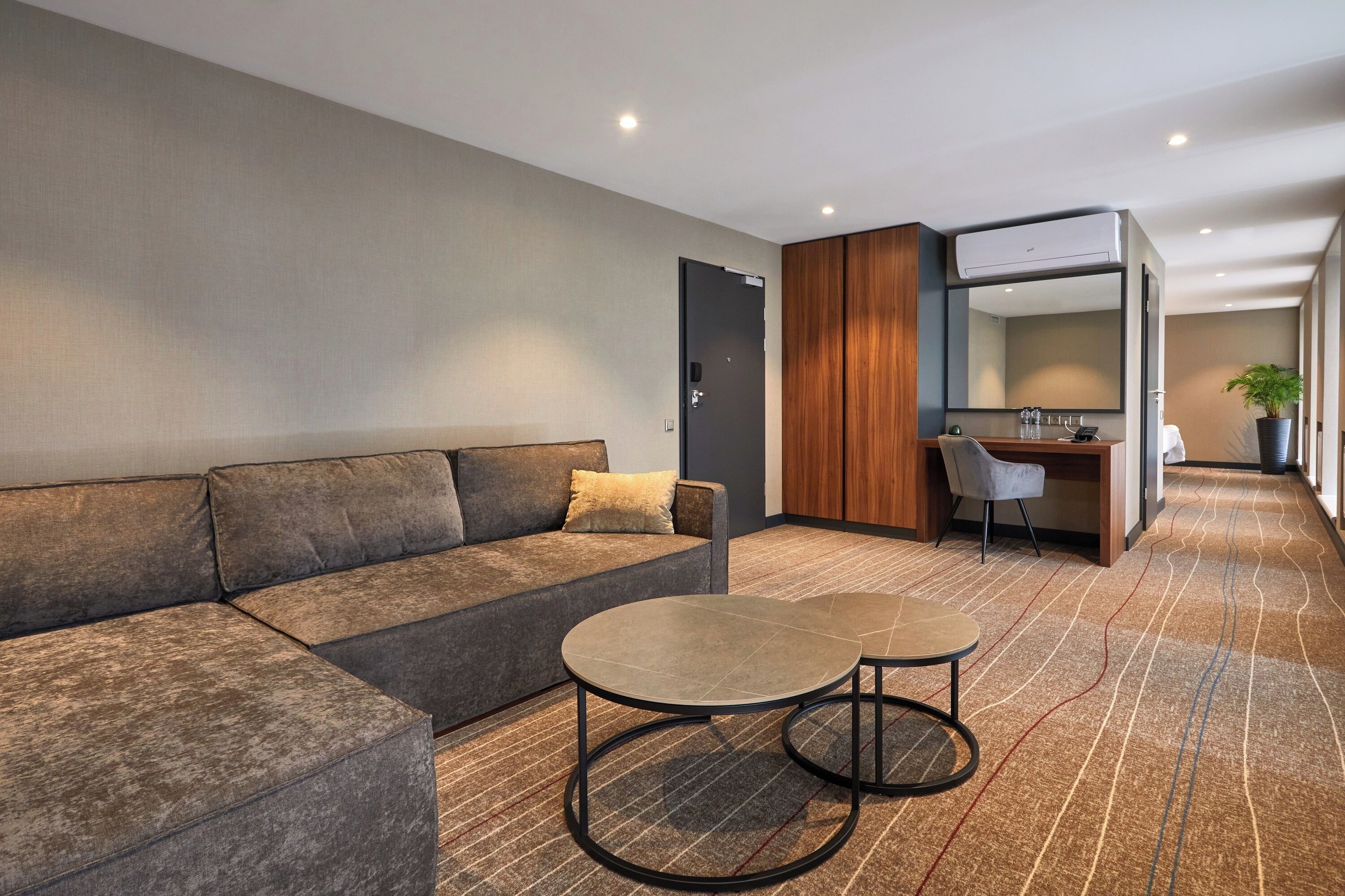 Una habitación de hotel moderna con un sofá modular, dos mesas redondas, un escritorio con una silla y un espejo grande.
