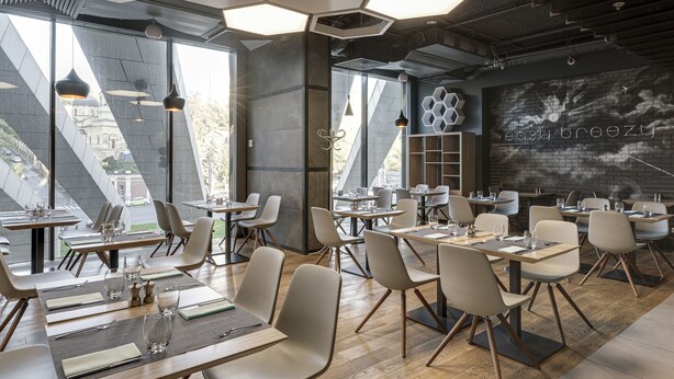A modern restaurant with hexagonal ceiling lights, a brick wall with a neon sign, and a view of a building through large windows. - Sky Loft Hotel by Rixwell (Kyiv)