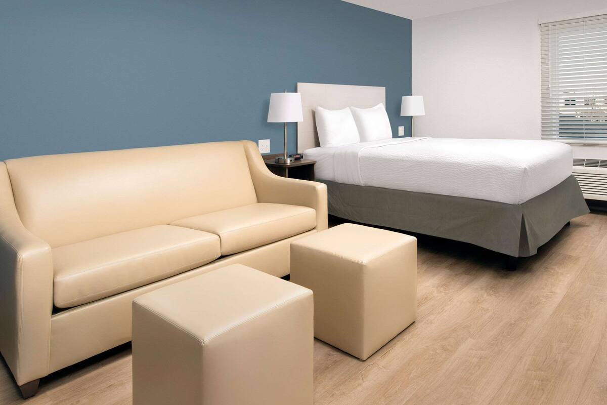 A hotel room with a beige sofa, a matching ottoman, a bed with white linens, and a nightstand with a lamp.