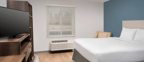 A modern hotel room with a bed, a desk with a flat-screen TV, a window with blinds, and a built-in air conditioning unit.