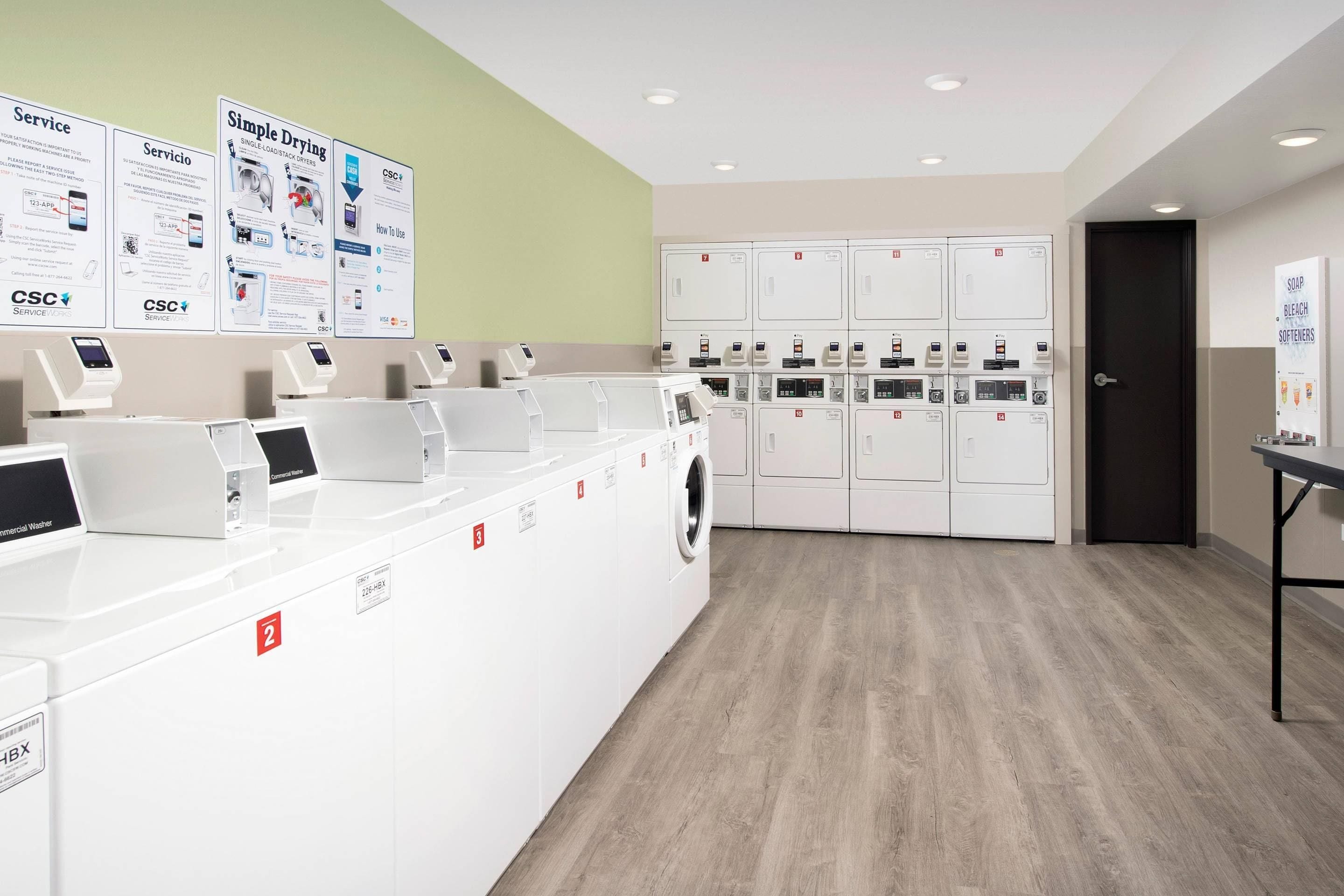 A commercial laundry facility with multiple washing machines and dryers, service posters on the wall, and a wooden floor.