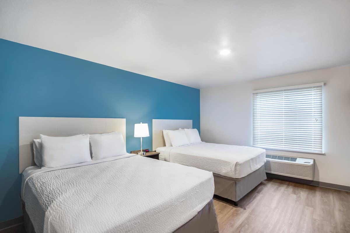 A hotel room with two beds, a blue accent wall, a window with blinds, and a built-in heater.