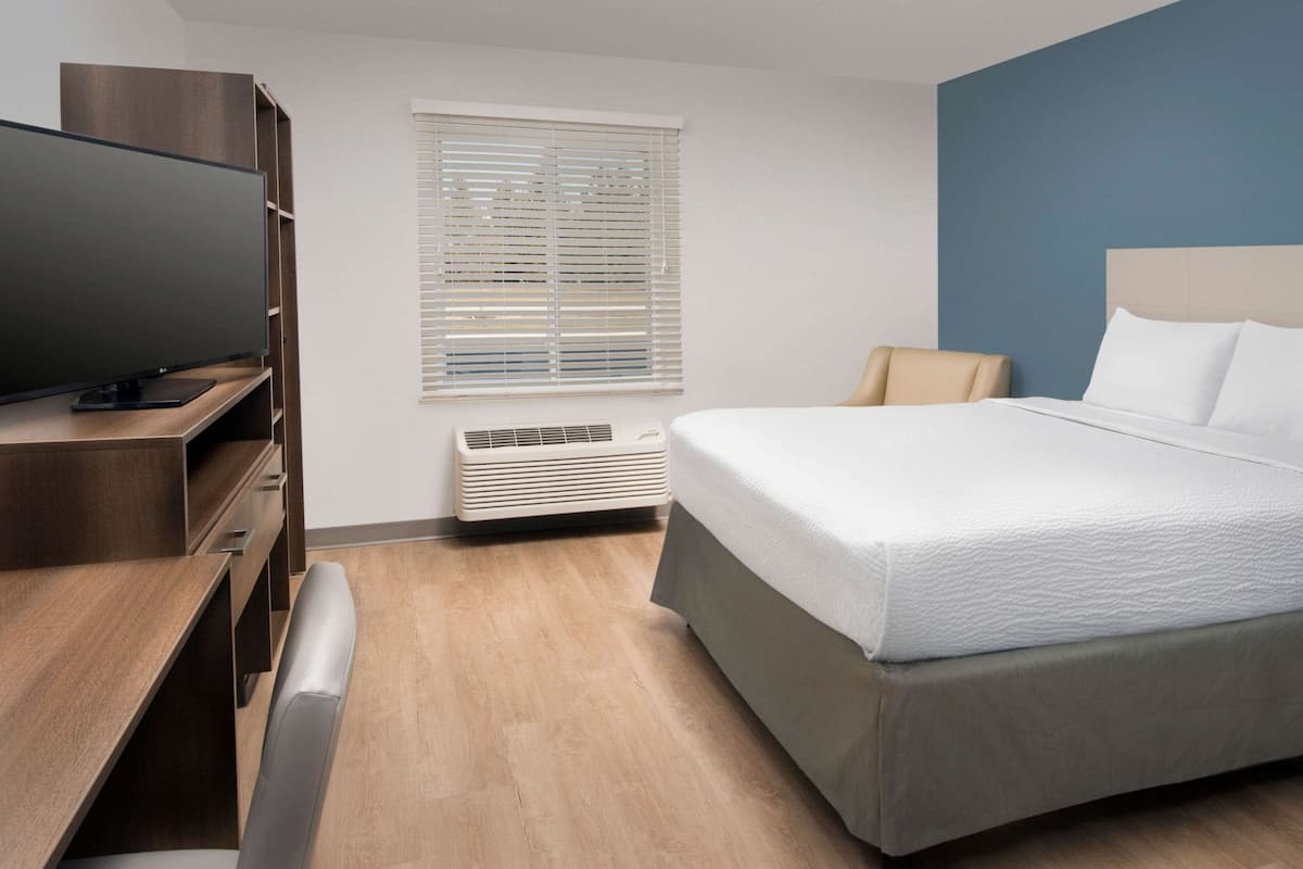 A modern hotel room with a wooden desk, a flat-screen TV, a bed with white linens, a beige chair, and a window with blinds.