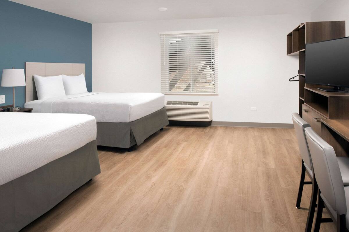 A hotel room with two beds, a desk with a chair, a television, a window with blinds, and an air conditioning unit.
