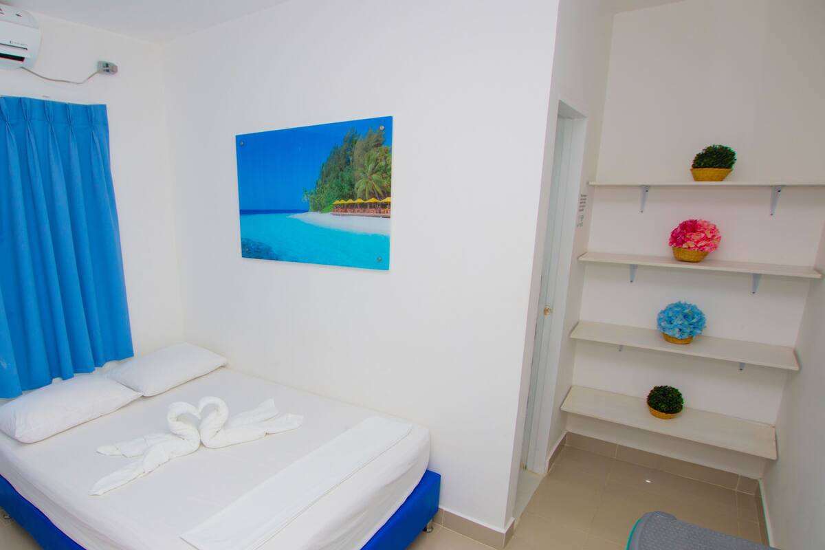 Een slaapkamer met een bed, een blauw gordijn, een luchtkoeler aan de muur, een foto van een strand en een plank met potplanten.