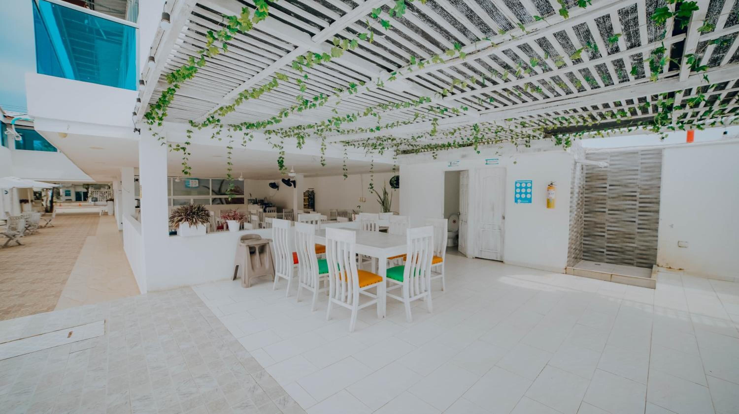 Un espacio interior amplio con una mesa y sillas de comedor, un techo con plantas colgantes y una pared exterior blanca.