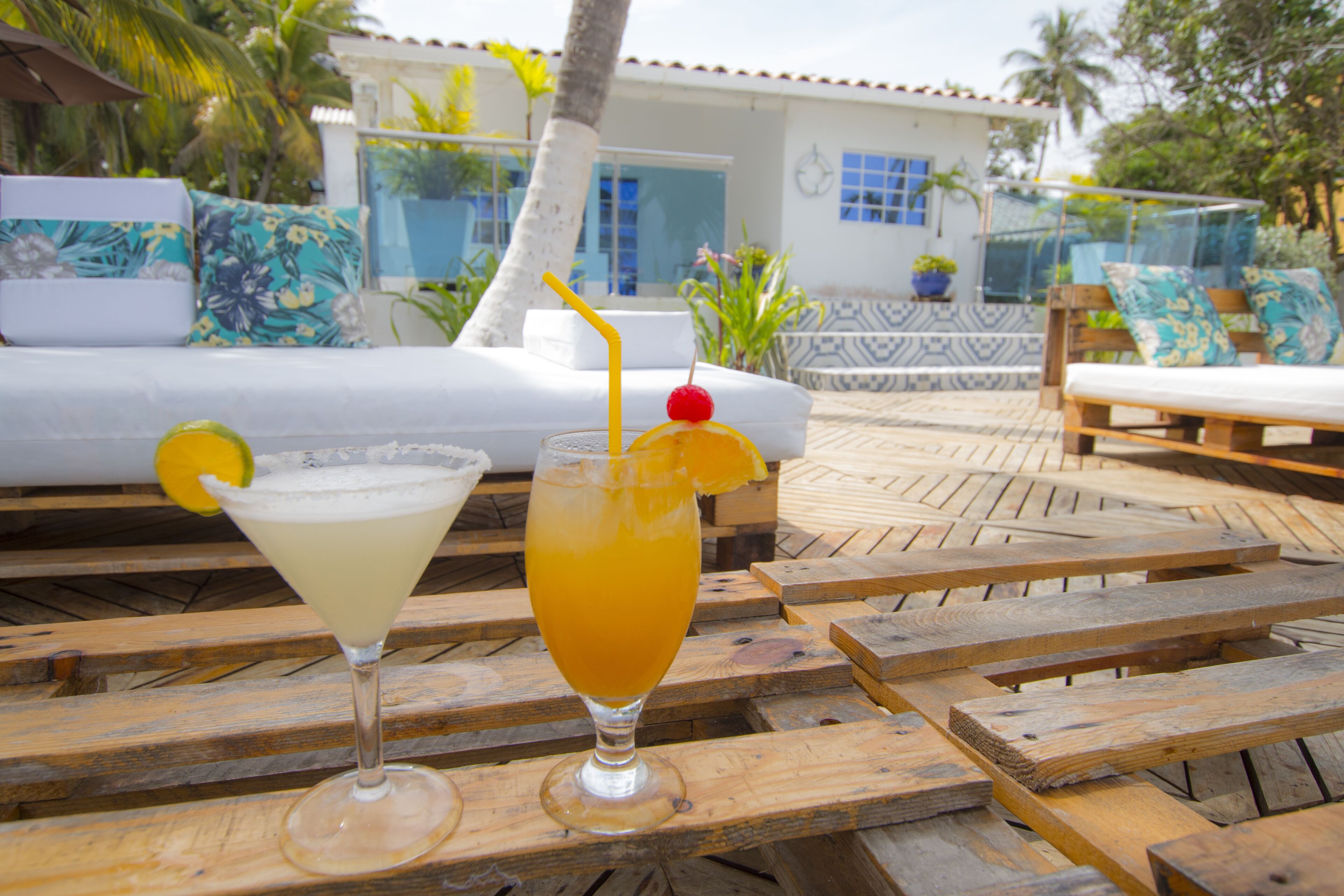 Una mesa de madera con dos bebidas, un sofá blanco y un patio con baldosas y palmeras al fondo.