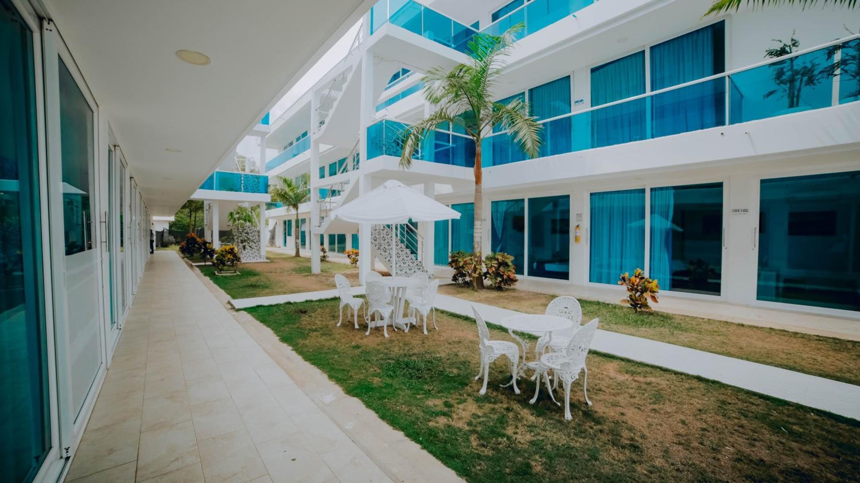 Un pasillo moderno de hotel con paredes exteriores blancas y azules, un área de estar exterior cubierta con sillas y una mesa blancas, y un pequeño jardín con una palmera.