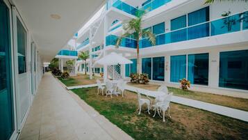 Un pasillo moderno de hotel con paredes exteriores blancas y azules, un área de estar exterior cubierta con sillas y una mesa blancas, y un pequeño jardín con una palmera.