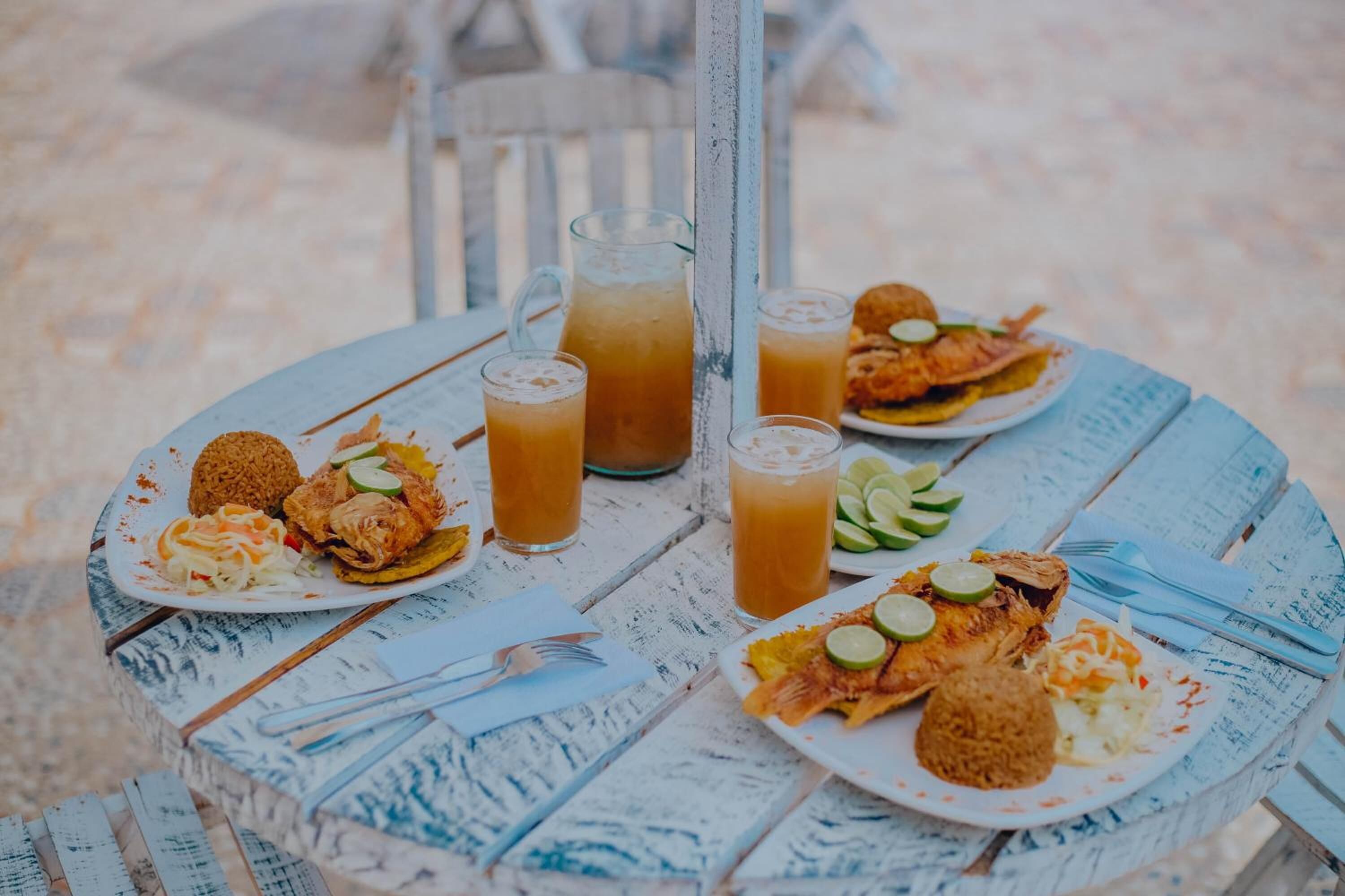Una mesa redonda con un acabado blanco y azul desgastado, rodeada de platos con comida y bebidas, incluyendo frituras y una jarrón de té helado.