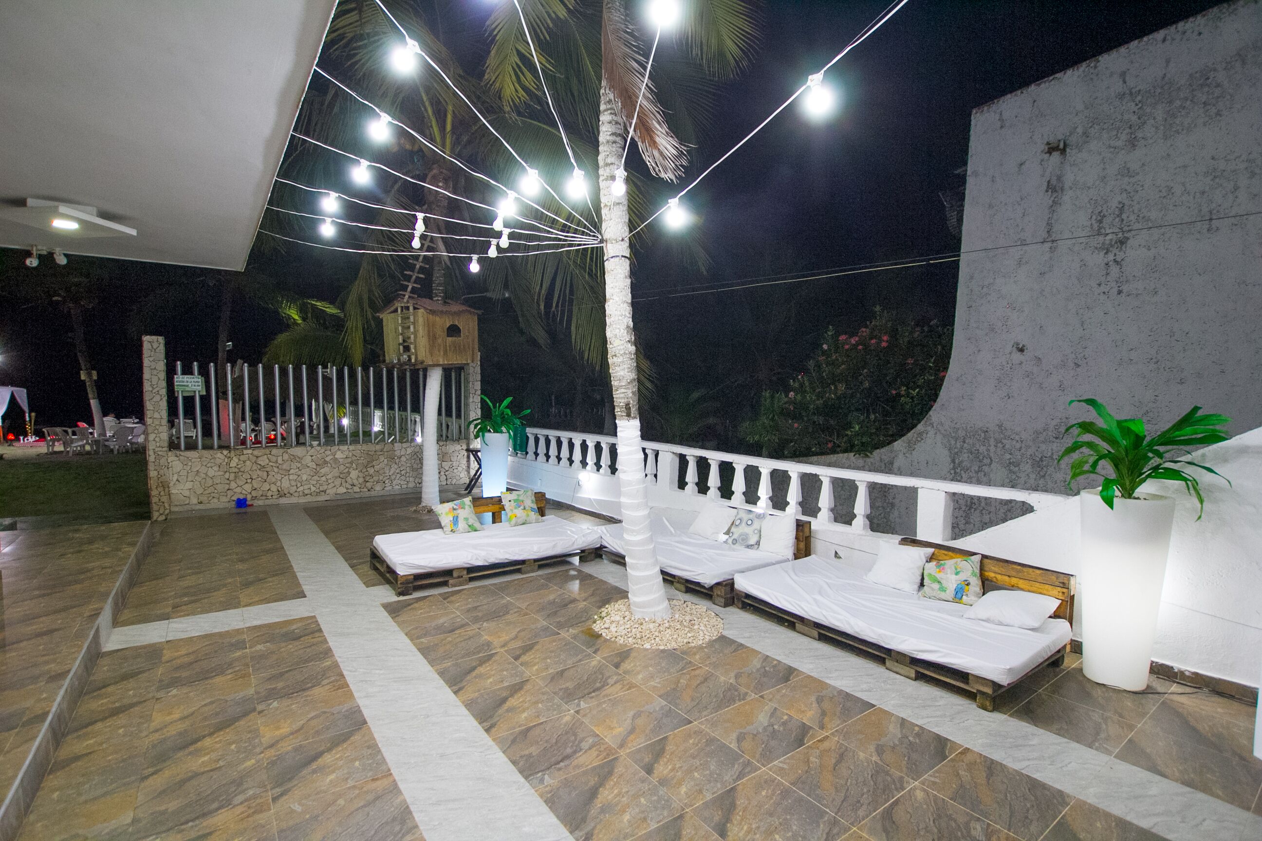 A rooftop terrace with a white railing, a stone wall, and a palm tree. There are two white sofas with cushions and a small table.