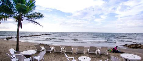 Un área de estar frente a la playa con sillas y mesas de plástico blanco, una palmera y el océano al fondo.