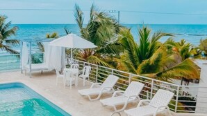 Área da piscina com espreguiçadeiras e uma mesa, com vista para o mar.