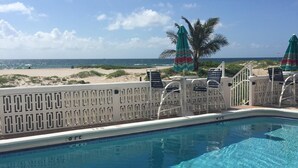 Une piscine à l’eau d’un bleu cristallin, entourée de chaises et de parasols blancs, avec vue sur une plage de sable et l’océan.