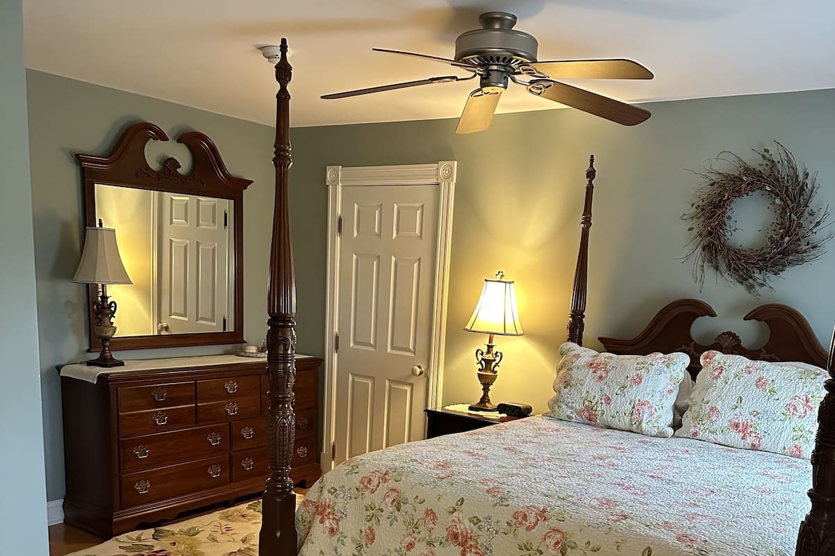 A bedroom with a four-poster bed, a ceiling fan, a dresser, a mirror, a door, and a wall wreath.