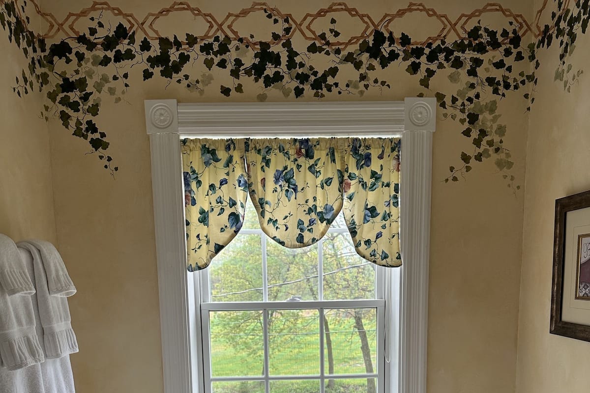 A bathroom with a large bathtub, a window with a floral valance, and a view of greenery outside.
