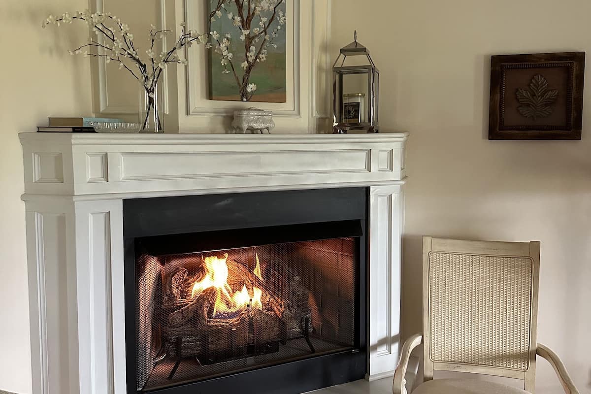 A fireplace with a fire, a white mantelpiece, a vase with branches, and a framed picture on the wall.