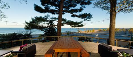 A wooden deck with a picnic table and wicker furniture, overlooking a body of water and a road.