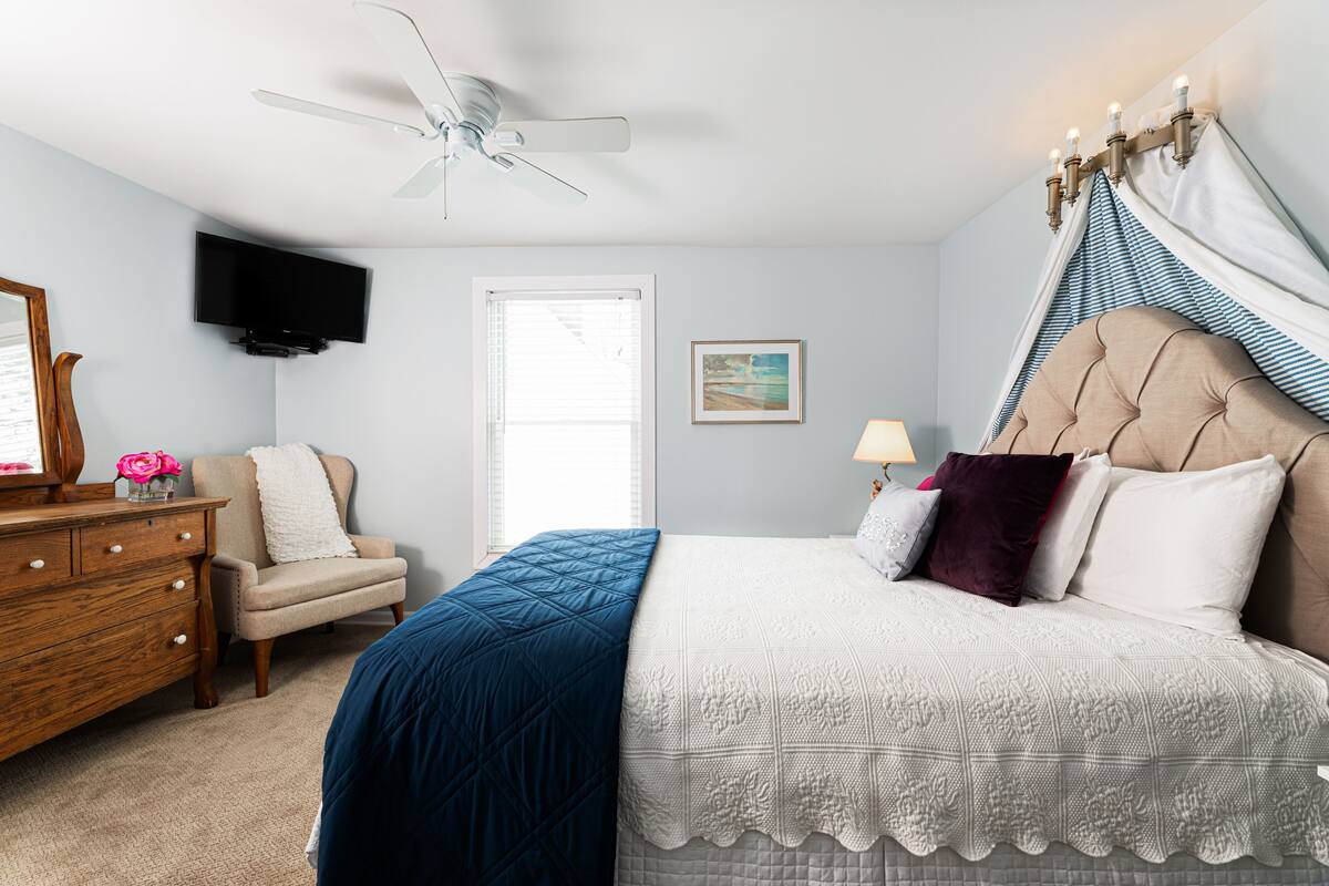 A bedroom with a bed, dresser, chair, TV, and a ceiling fan.
