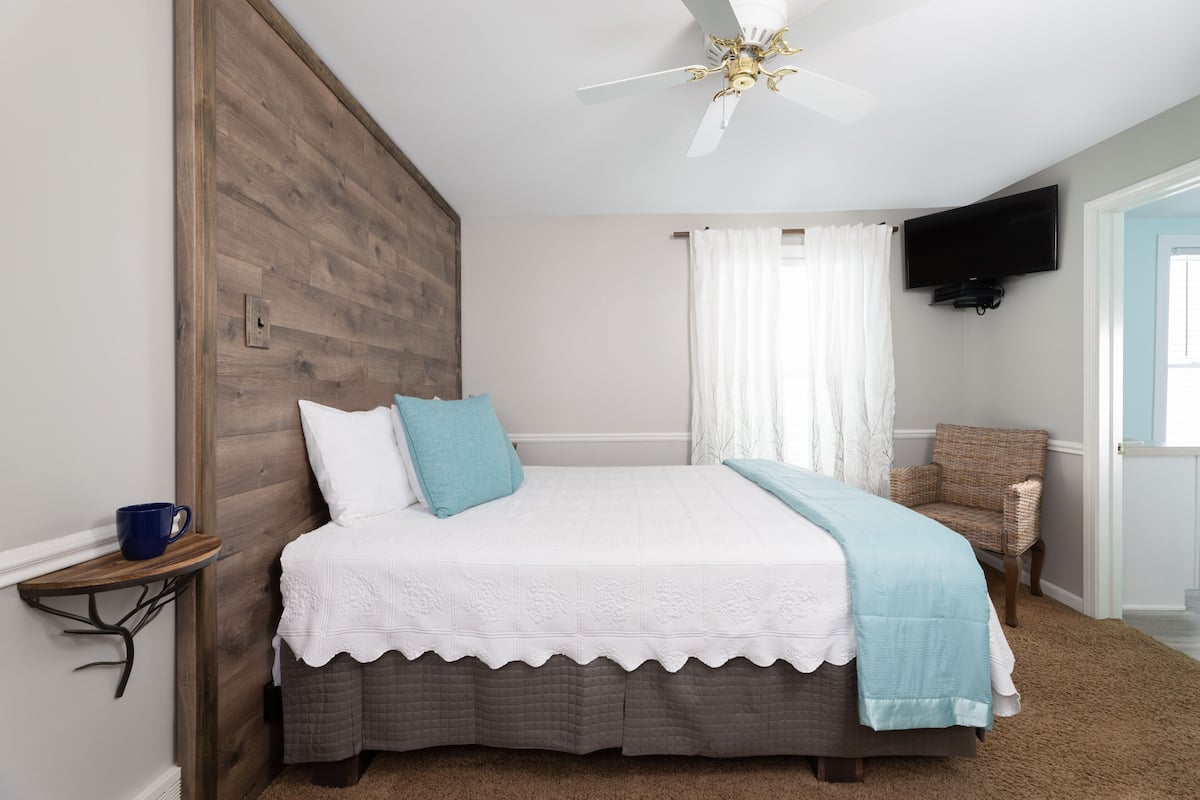 A bedroom with a bed, a ceiling fan, a television, a chair, and a window with curtains.