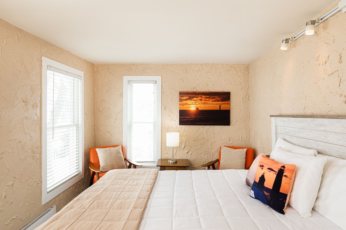 A bedroom with a large bed, two chairs, a nightstand, a lamp, and a painting of a sunset.