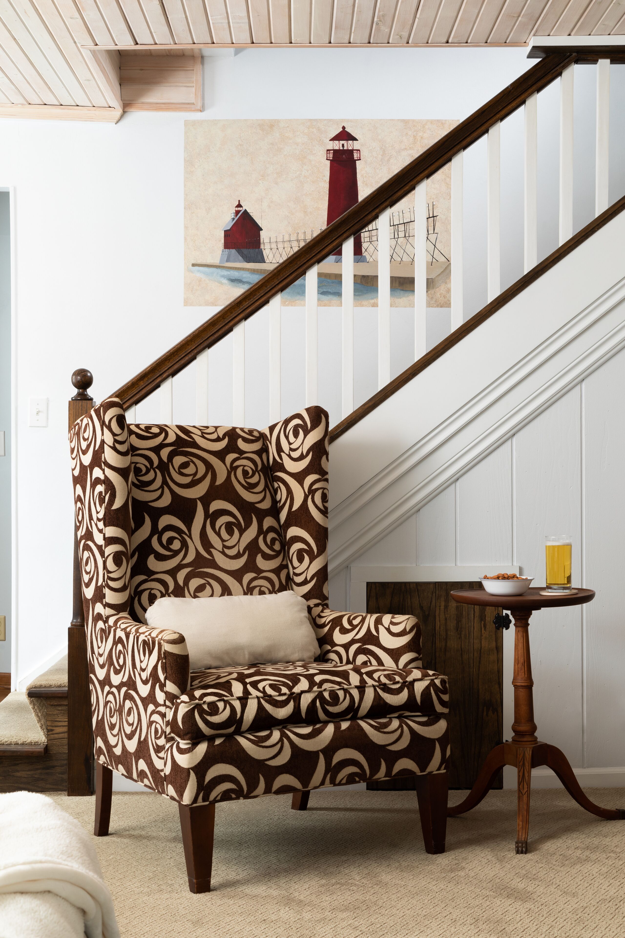 Una silla con un diseño estampado, una mesa de madera con un vaso, y un cuadro de un faro en la pared.