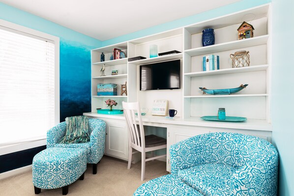 Una habitación con un escritorio blanco, dos sillas con estampado, una estantería y una ventana con persianas.