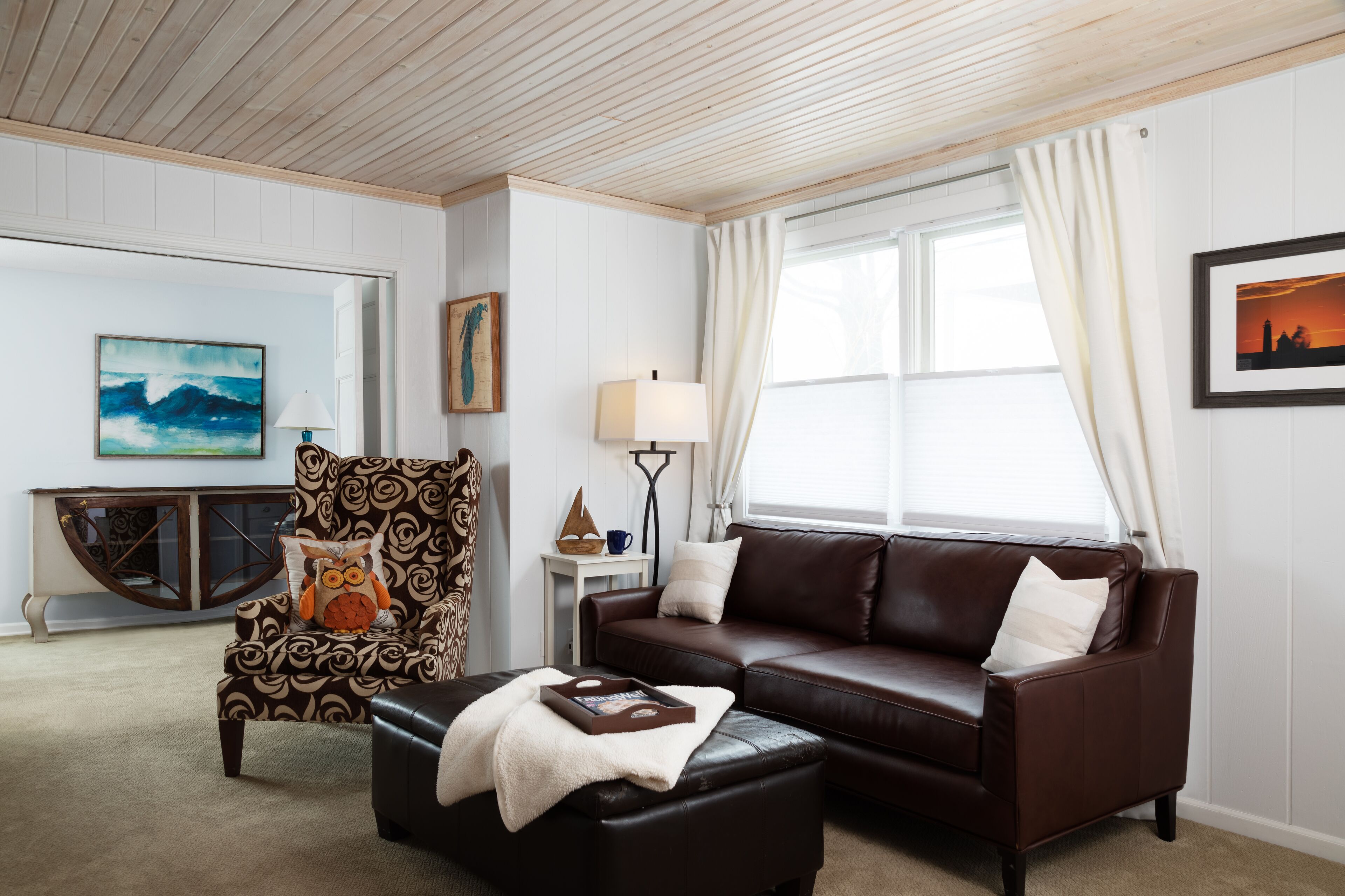 A living room with a brown leather sofa, a patterned armchair, and a black ottoman. There is a lamp on a side table, a painting on the wall, and a window with curtains.