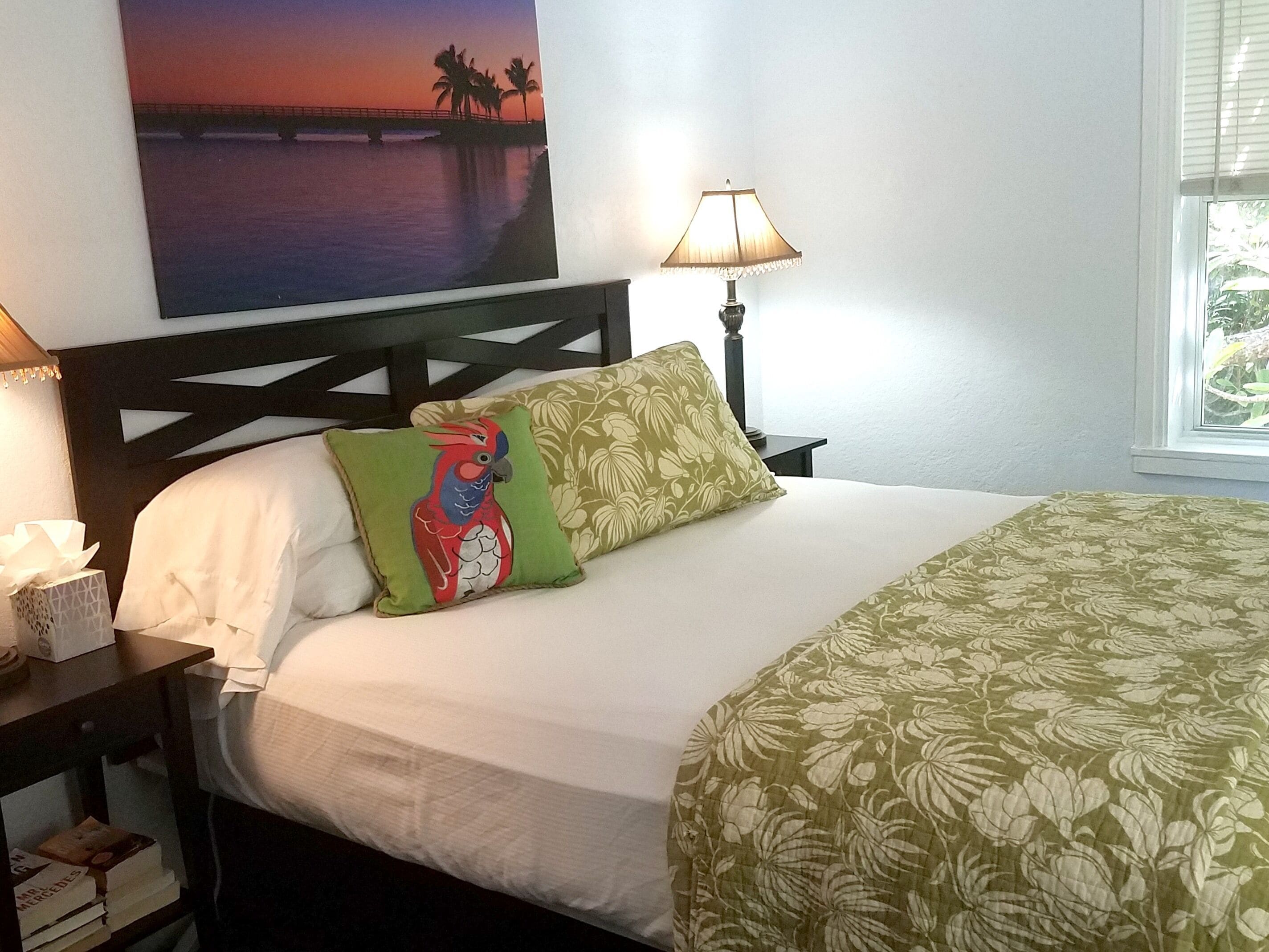 A neatly made bed with a white comforter and green floral bedspread, a green and red parrot pillow, a lamp on a bedside table, and a painting of a sunset over water on the wall.