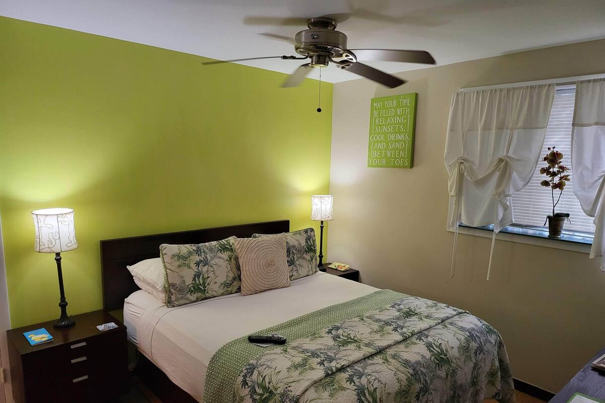A bedroom with a bed, nightstands, a ceiling fan, a window with curtains, and a wall with a motivational quote.