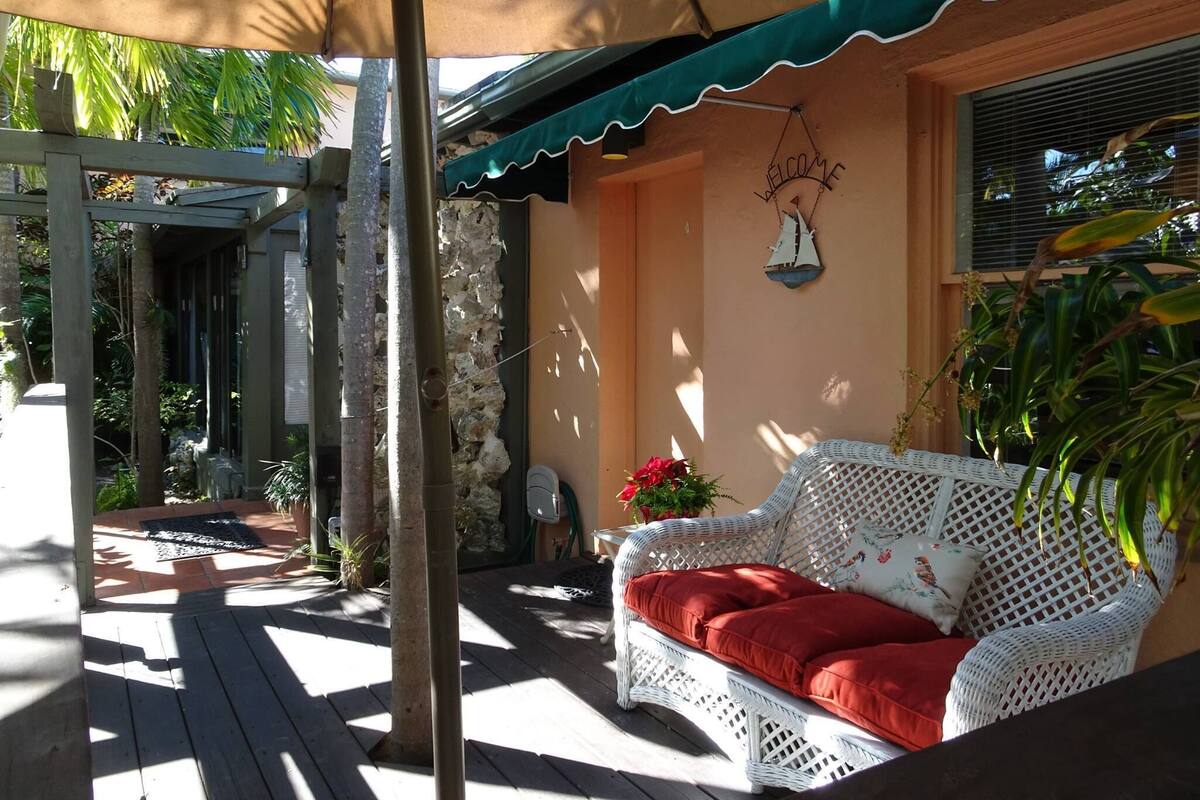 A covered outdoor seating area with a white wicker sofa, a red cushion, and a 'WELCOME' sign.