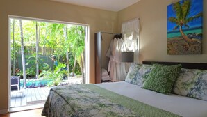 Un dormitorio con una cama grande, vista a un jardín tropical a través de una puerta corredera, y un cuadro de una palmera en la pared.