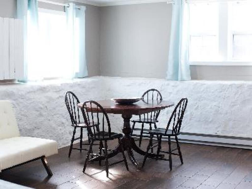 A dining area with a round table and four chairs, a white sofa, and a window with light blue curtains.