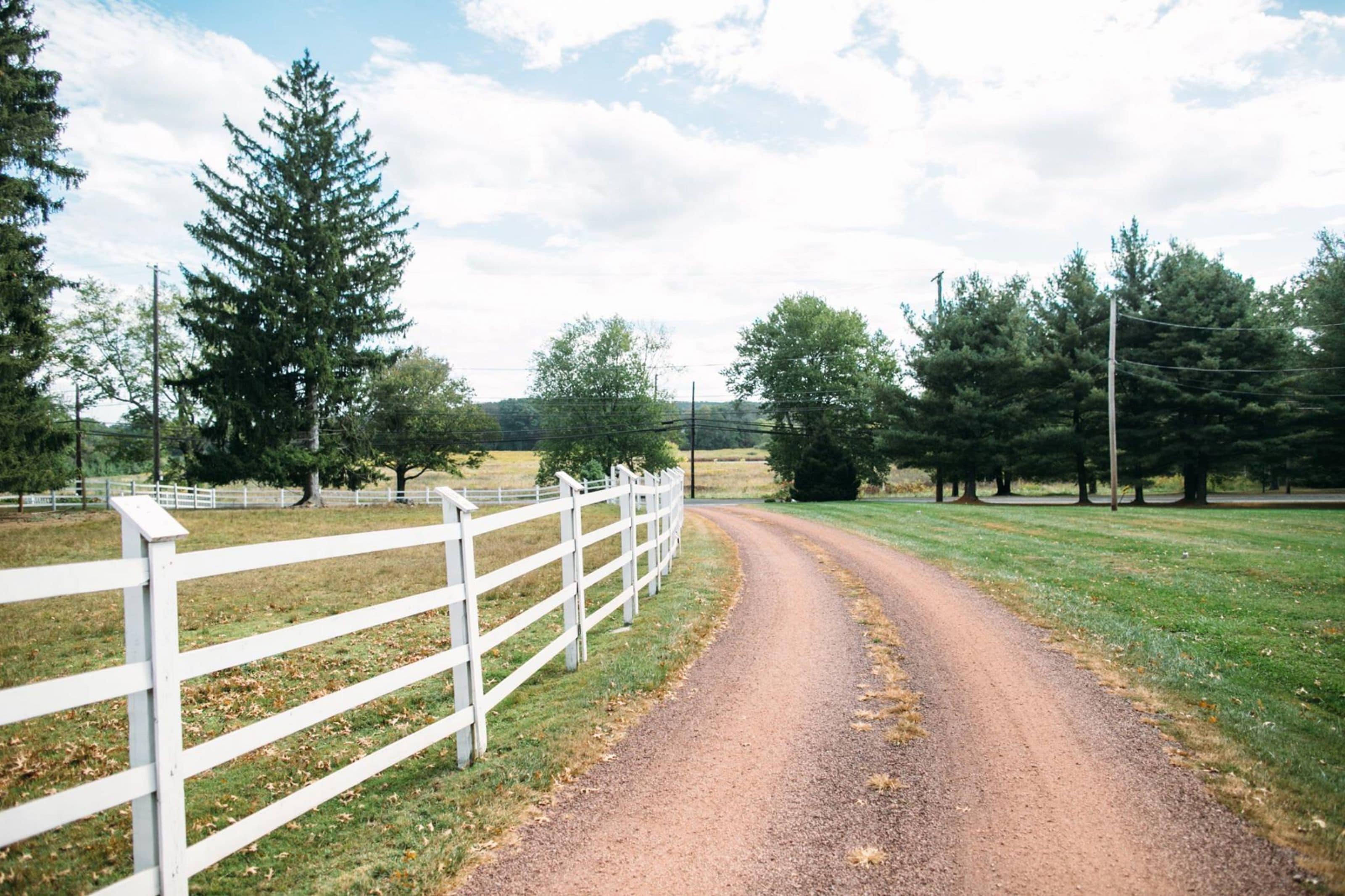 A rural dirt road with a white fence on one side and trees on the other.