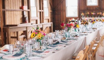 A long dining table set with glassware, plates, and colorful floral centerpieces in a rustic wooden room.