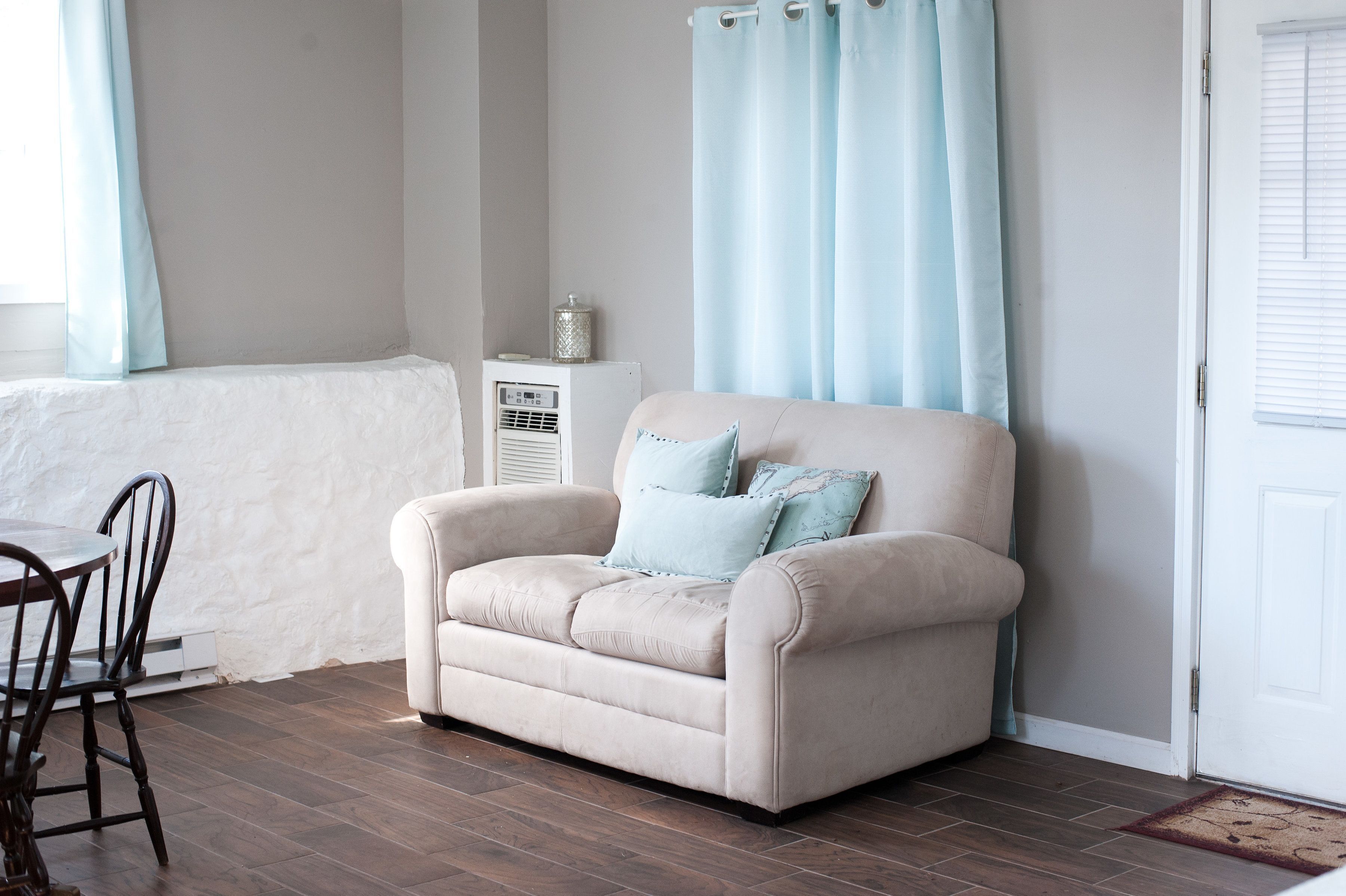 Un canapé beige agrémenté de deux coussins bleu clair, un radiateur blanc, une table à manger en bois avec des chaises, et une porte blanche avec une fenêtre.