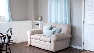 Un sofá beige con dos almohadas azul claro, un radiador blanco, una mesa de comedor de madera con sillas y una puerta blanca con ventana.