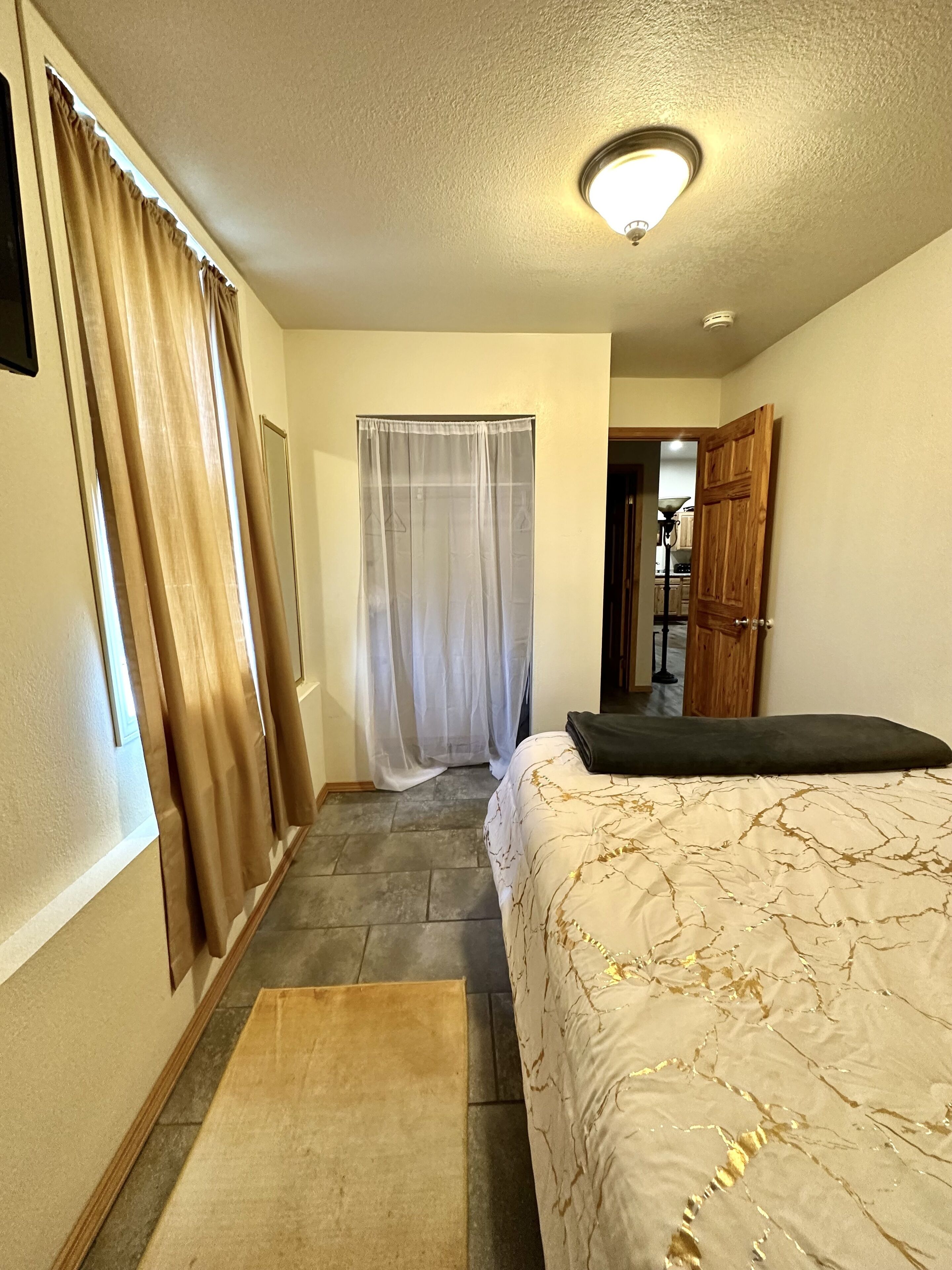 Une chambre d’hôtel avec un lit, une porte en bois, une fenêtre avec des rideaux et un couloir menant à une autre pièce.
