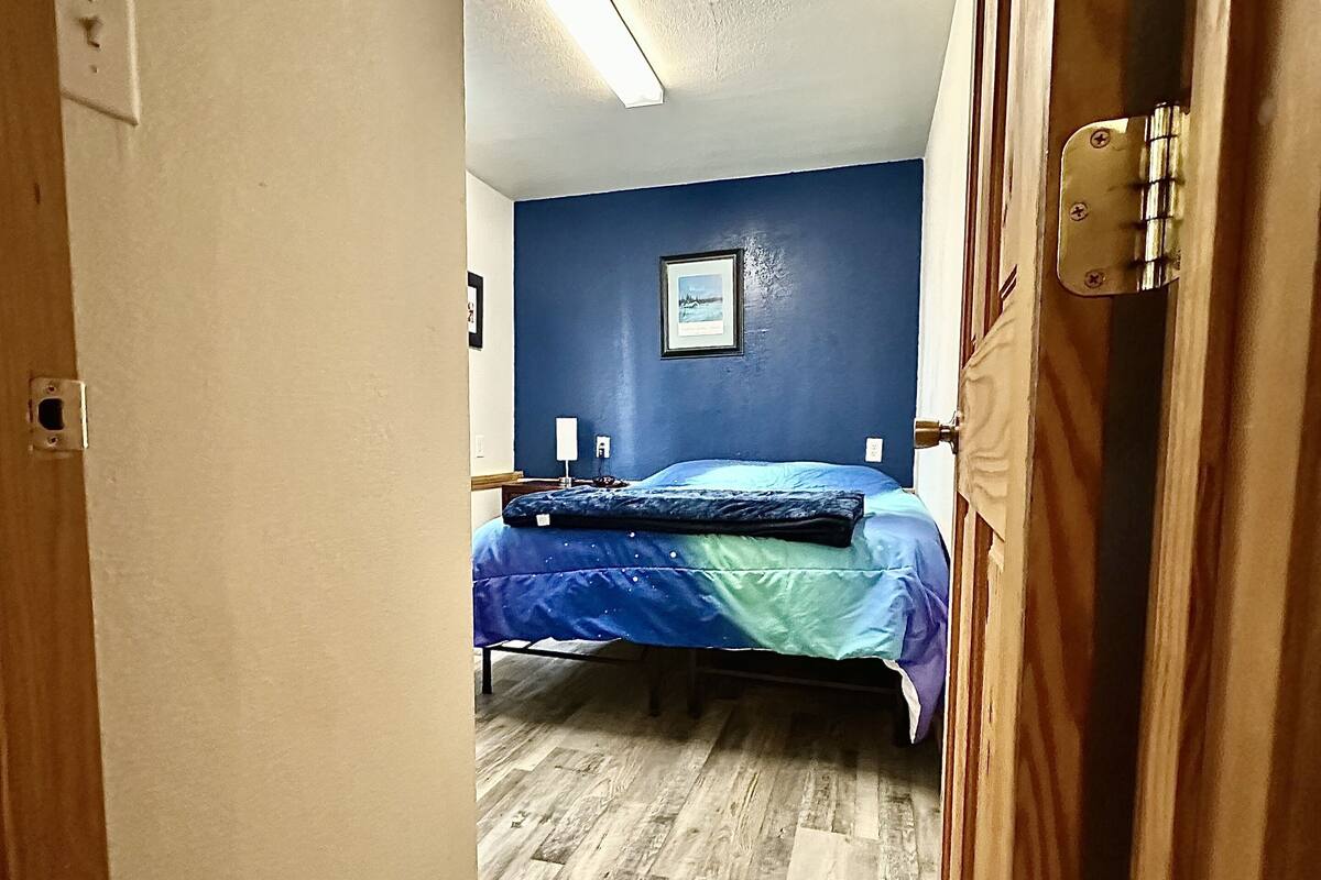 Kapea käytävä johtaa makuuhuoneeseen, jossa on sänky, seinällä kehystetty kuva ja puulattia.