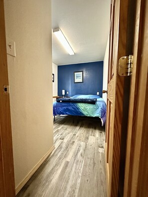 Un couloir étroit mène à une chambre à coucher qui contient un lit, un tableau encadré accroché au mur et un plancher en bois.