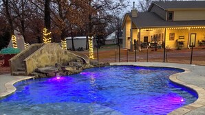 Una piscina con illuminazione blu, una cascata di pietra e una casa sullo sfondo con luci a catena.