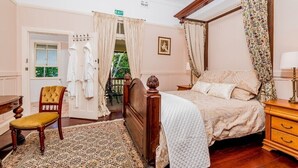 A four-poster canopy bed with a quilted bedspread, a wooden nightstand, a chair with a cushion, a patterned rug, and a door with an exit sign.