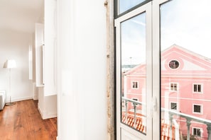 A room with a wooden floor, white walls, and a large window overlooking a pink building with a balcony. - The Lisbon-Way Apartments (Lisbon)