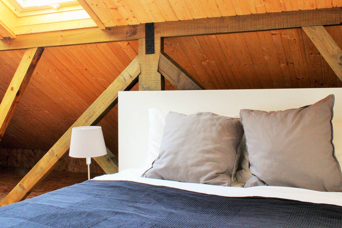 Un dormitorio con techo inclinado, vigas de madera, una cama con dos almohadas y una lámpara de noche.